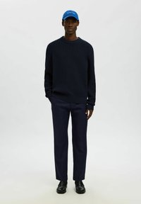 Uomo che indossa un maglione blu a maglia, pantaloni scuri abbinati, mocassini neri e un cappellino blu brillante con testo bianco, in piedi su uno sfondo neutro.
