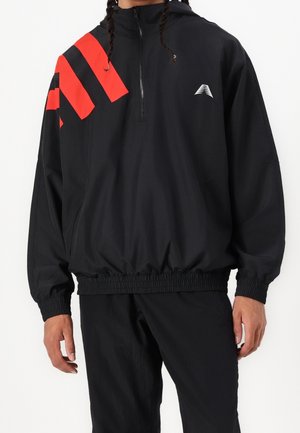 Mand iført en sort pullover vindjakke med røde geometriske striber på venstre skulder og elastiske manchetter og kant.
