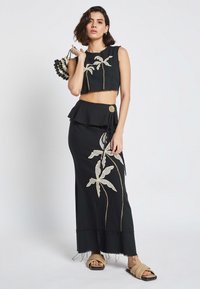 Completo nero in due pezzi composto da un top corto con ricami di foglie di palma e una lunga gonna con vita peplum. Include un bottone decorativo.