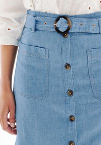 Jeanskjol i ljusblå nyans, med ett bälte i midjan som har en stor rund spänne, två framfickor och bruna knappar längs framsidan.