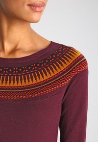 Pull bordeaux avec un empiècement orné de motifs géométriques jaunes, rouges et noirs. Tissu doux avec un col rond.