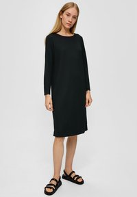 Schwarzes langärmeliges Kleid mit Rundhalsausschnitt, strukturiertem Stoff und seitlichen Schlitzen, kombiniert mit schwarzen Sandalen mit Riemen auf einem neutralen Hintergrund.