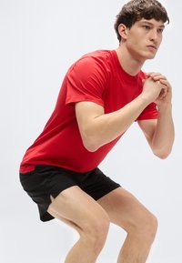 Κόκκινη βαμβακερή μπλούζα συνδυασμένη με μαύρο αθλητικό σορτς. Το μοντέλο βρίσκεται σε θέση καθίσματος, παρουσιάζοντας το στρογγυλό γιακά και τα κοντά μανίκια της μπλούζας.