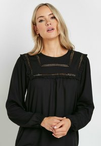 Blusa nera con maniche lunghe, caratterizzata da un colletto arricciato e dettagli geometrici intagliati intricati. Vestibilità comoda, texture morbida.