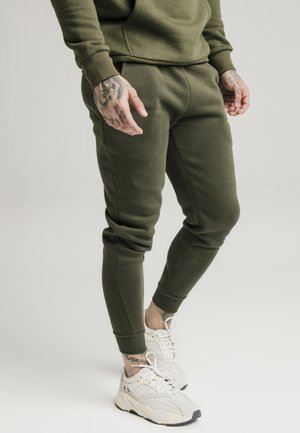 Mann trägt olivgrüne Jogginghose und Sweatshirt mit weißen Sneakers, zeigt tätowierte Hände und Knöchel, steht vor einfachem Hintergrund.