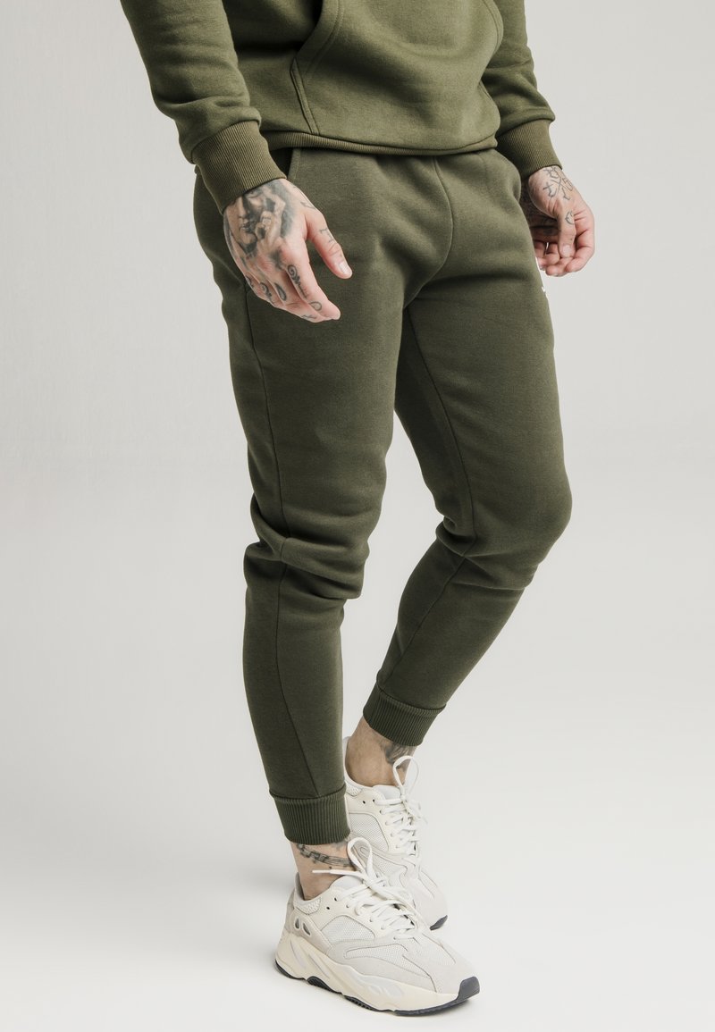 Homme portant un pantalon de survêtement vert olive et un sweat-shirt avec des baskets blanches, montrant des mains et des chevilles tatouées, debout sur un fond uni.