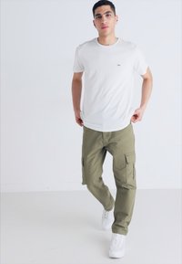 Fehér pamut póló kis logóval, zöld olajzöld cargo nadrággal, oldalzsebekkel és fehér sportcipővel egy egyszínű háttéren.