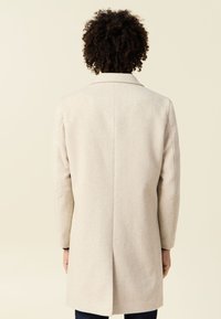 Abrigo de lana color beige claro con una abertura trasera, forma ajustada y cuello de solapa estándar. Textura suave y líneas limpias.