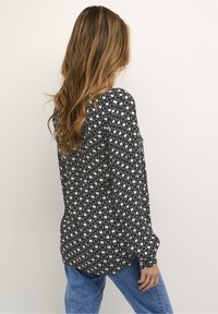 Kobieta z długimi falującymi włosami, ubrana w wzorzystą czarno-białą bluzkę i niebieskie dżinsy, stojąca na tle jednolitego białego tła.