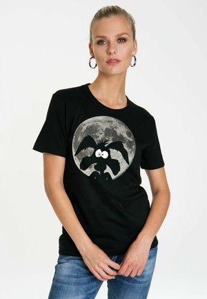Woman with light hair tied back wears black t-shirt with cartoon dog silhouette and full moon graphic, paired with blue jeans.