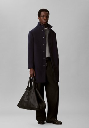 Mann trägt einen dunkelblauen Mantel, grauen Pullover, schwarze weite Hosen und schwarze Schuhe, hält eine große schwarze Leder-Tasche.