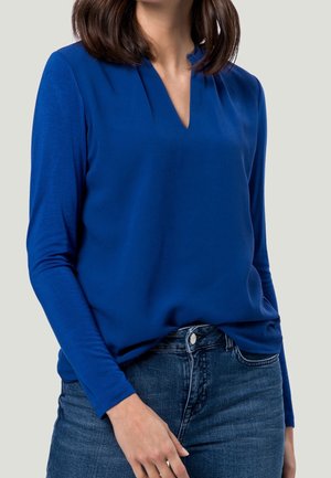 Blue long-sleeve top with a V-neck design and soft texture, paired with dark denim jeans, featuring a fitted silhouette and minimal details.