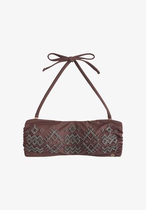 Vrouw in een bruin bikini met patroon en een zwarte hoofdband staat met opgetilde armen een tak van een boom vasthoudend in een buitenomgeving met een houten hek.