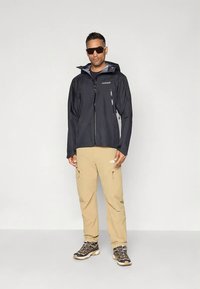 Zwarte waterdichte jas met capuchon en ritszakken, gecombineerd met beige broek en bruine wandelschoenen. Donkere zonnebril maakt de look compleet.