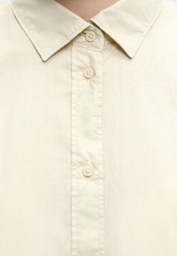 Κοντινό πλάνο για το γιακά πουκάμισου σε κρεμ απόχρωση με τρία αντίστοιχα κουμπιά κλεισμένα πάνω σε λείο ύφασμα.