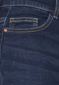 Vaqueros de mezclilla azul oscuro con un pequeño bolsillo, costuras amarillas en contraste y un detalle de remache de cobre en la parte delantera.