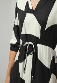 Czarna i biała sukienka w geometryczny wzór z dekoltem w kształcie litery V, rękawami trzy czwarte i regulowanym paskiem w talii na torsie osoby.