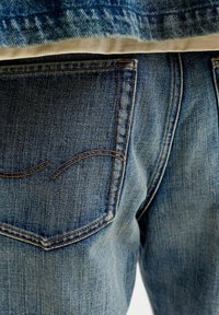 Bagsiden af blå cowboybukser med synlige syninger på baglommen og en del af en linning med en hvid skjorte stukket ind.