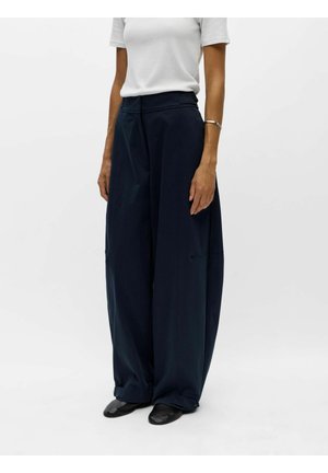 Personne portant un pantalon large bleu marine, un haut blanc à manches courtes, des chaussures noires et un bracelet argenté au poignet droit sur un fond uni.