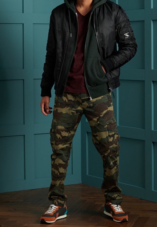 Sort bomberjakke lagdelt over en grøn hættetrøje, parret med camo cargo bukser og sneakers med orange accenter. Trægulv og teal væg som baggrund.