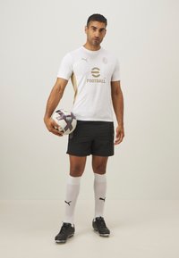 Maglia da calcio bianca con dettagli dorati, pantaloni corti neri, calzettoni bianchi, che tiene in mano un pallone da calcio. Presenta il marchio Puma e un design in tessuto testurizzato.