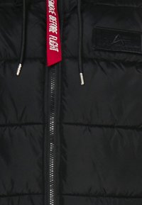 Jachetă puffer neagră cu un fermoar central, textură matlasată și etichetă roșie "REMOVE BEFORE FLIGHT" cu accente argintii.