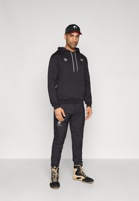 Casaco com capuz preto com bolso frontal, combinado com calças de treino pretas com padrão e sapatos desportivos bege. Boné preto com detalhes em logo.