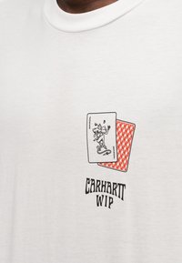 Λευκό κοντομάνικο t-shirt με γραφικό δύο παιγνιδιάρικων φύλλων, το ένα κόκκινο με γεωμετρικό σχέδιο, και το κείμενο "CARHARTT WIP" από κάτω.