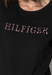 Schwarzes Baumwollo T-Shirt mit "HILFIGER" in weißen und roten, blumenmusternden Buchstaben. Rundhals und kurze Ärmel.
