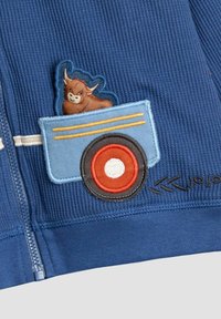 Tessuto a coste blu con una toppa ricamata che raffigura un animale marrone in un carrello blu con una ruota rossa e bianca, accentuato da linee gialle.