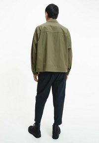 Olivgrüne Jacke mit Kragen, langen Ärmeln und lockerer Passform, kombiniert mit dunklen, schmal zulaufenden Hosen und schwarzen Schnürstiefeln.