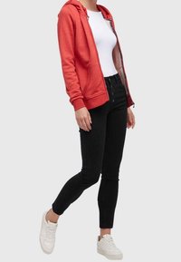 Rød zip-up hoodie med forlommer, båret over en hvid shirt og parret med sorte skinny jeans og hvide sneakers. Glat stof tekstur.