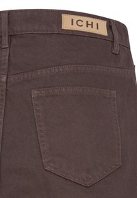 Gros plan d'une poche arrière d'un pantalon en denim marron avec passants de ceinture et étiquette beige indiquant "ICHI" sur la ceinture.