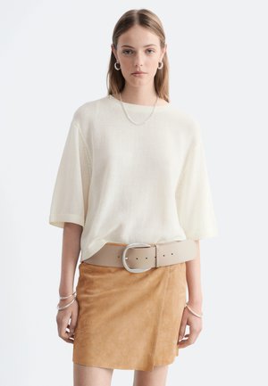 Jeune femme portant un pull blanc ample à manches courtes, une jupe en daim beige avec une large ceinture beige, ainsi que des créoles et des bracelets en argent.