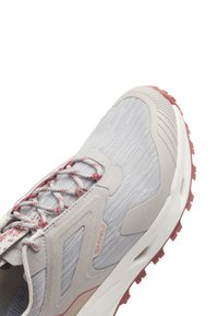 Chaussure de sport gris clair avec tige en tissu texturé, lacets roses et semelle accentuée. Présente un logo et un design ondulé sur le côté.