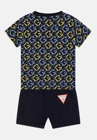 Košulja kratkih rukava tamnoplave boje s geometrijskim uzorkom u žutoj i svijetloplavoj boji, u kombinaciji s mornarskim šortsevima koji imaju crvenu i bijelu logotip oznaku.