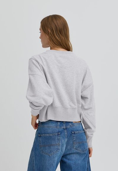 Sivý skrátený mikina s textúrovaným povrchom, širokými rukávmi a rebrovaným lemom, skombinovaná s modrými džínsami s vysokým pásom, ktoré majú zadné vrecká.