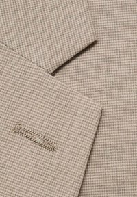Beige texturerad tyg med ett litet rutmönster, med en sömdetalj och subtila tonvariationer över ytan.