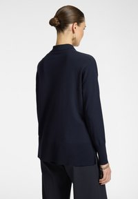 Pullover navy, in maglia leggera, design colletto, maniche lunghe, orlo a coste, spacchi laterali, indossato sopra pantaloni scuri a fantasia.