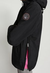 Schwarze Kapuzenjacke mit Reißverschluss vorne, Seitentaschen und einem runden Logoaufnäher am Ärmel. Das Innenfutter ist kontrastreich in Pink gehalten.