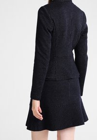 Laivastonsininen puuvillainen bleiseri ja sitä vastaava kellohame. Sileä kangas ja vartalonmyötäinen siluetti, jossa on pepitaraitainen yksityiskohta takana.