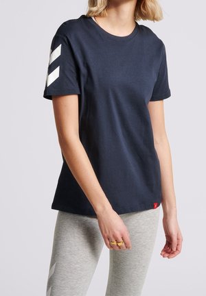 Kobieta ubrana w granatowy t-shirt z krótkim rękawem, z białymi paskami w kształcie jodełki na rękawie oraz jasnoszare legginsy z pasującymi akcentami w jodełkę.