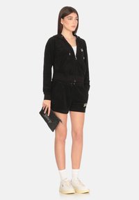 Completo in velluto nero composto da una felpa con zip e pantaloni shorts, abbinato a calzini bianchi e sneakers beige, mentre tiene una borsa a pochette nera.