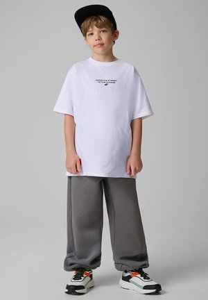Chłopiec w białej oversize koszulce, szarych szerokich spodniach, wielokolorowych butach sportowych i czarnej czapce, stojący na jednolitym szarym tle.