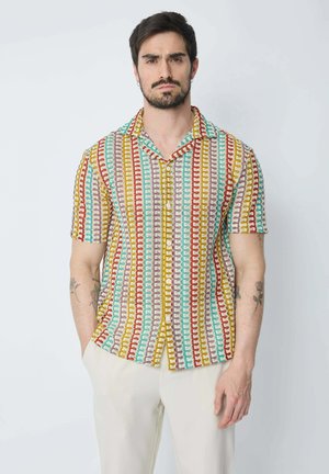 Homme aux cheveux foncés et à la barbe portant une chemise à manches courtes boutonnée avec des rayures verticales multicolores et un pantalon clair.