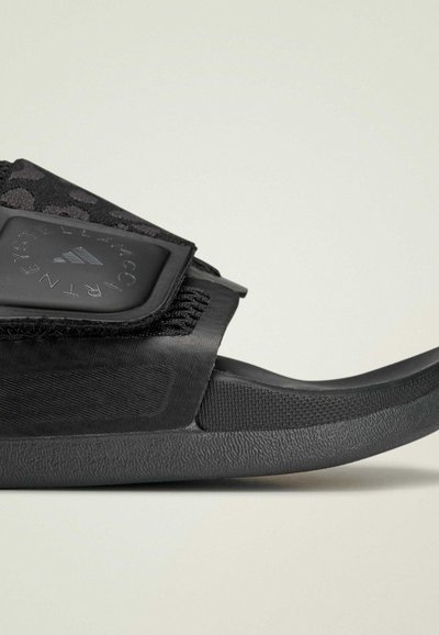 Sandale noire à glissière avec une bride texturée, comportant un patch logo en plastique. La semelle intérieure est rembourrée et la semelle extérieure possède un motif antidérapant.