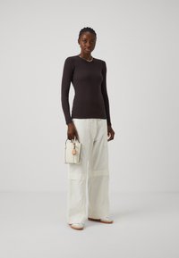 Haut marron à manches longues associé à un pantalon large blanc, tenant un petit sac à main blanc avec des accents noirs et une étiquette rose.