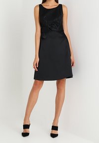 Czarna sukienka koktajlowa z dopasowaną górą, teksturowaną częścią górną oraz gładką spódnicą. Posiada detal kokardy w talii oraz bezrękawowy krój.