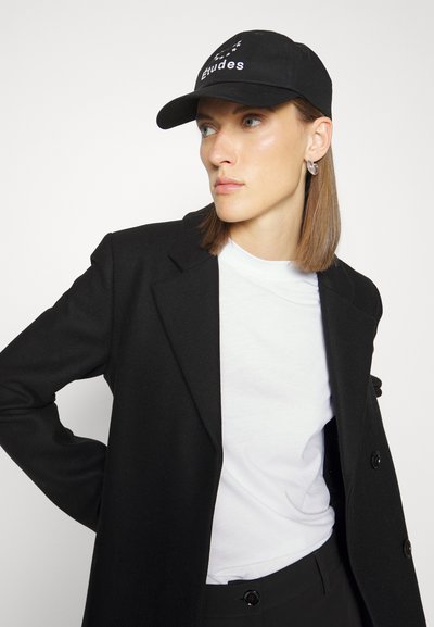 Black baseball cap with white embroidered text, worn with a black coat and white t-shirt, featuring a simple, classic design.