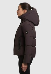 Korte pufferjas in donkerbruin, met een ruime capuchon, zwarte ritszak en ribgebreide manchetten voor warmte en comfort.